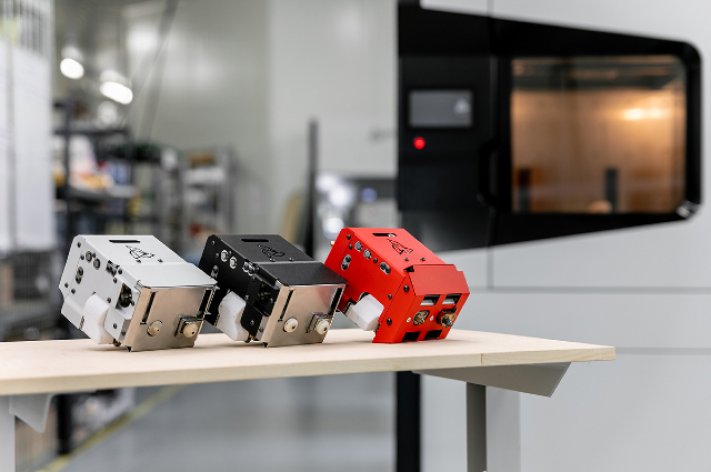 Trzy wymienne moduły drukujące M280 (biały(, M360 (czarny) i M500 (czerwony) leżą na stole. Za nimi stoi przemysłowa drukarka 3D - 3DGence INDUSTRY F420